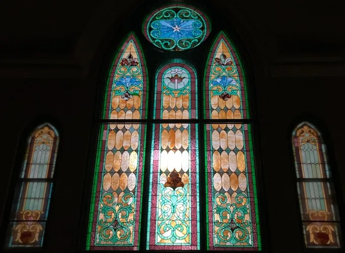 The north stained-glass windows lit up in the sanctuary: three tall arched panels in amber, green and blue beneath a round window with a star, between two smaller arched windows